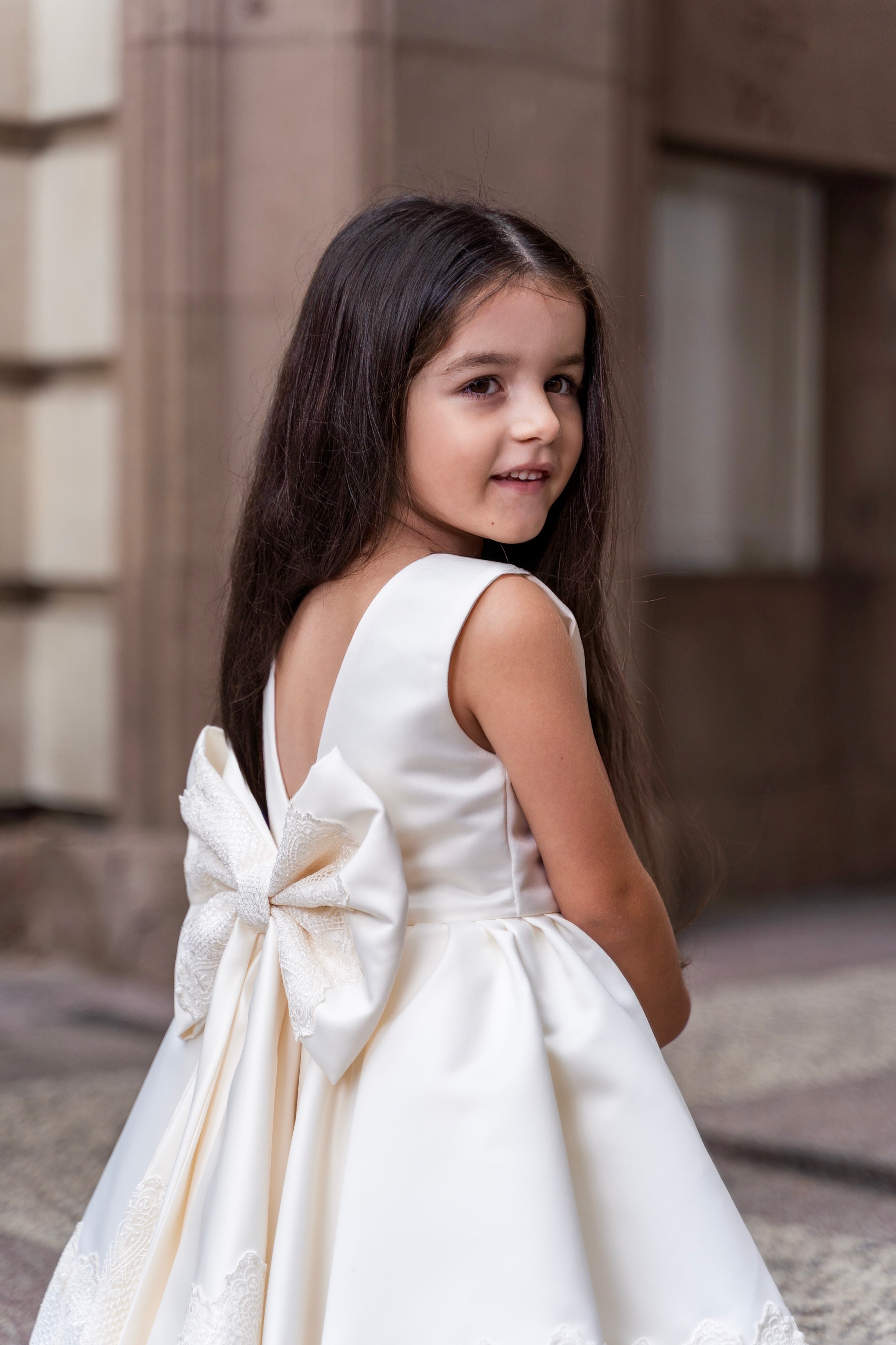 Young girl in a white satin dress with a large bow at the back, standing outdoors.
